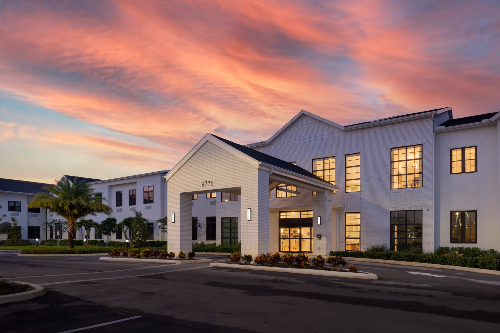 Exterior view of American House Boynton Beach facility at sunset, showing a white brick building with large windows, a covered entrance with the number 9776, landscaped plants, and a palm tree in the parking area.
