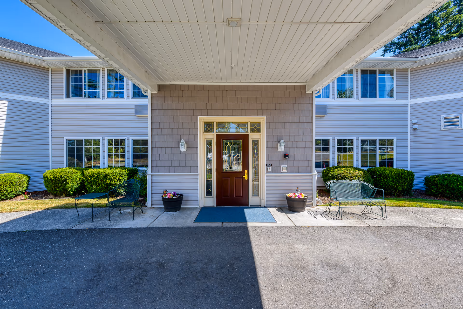 Entrance of a senior living facility with a covered driveway, a brown door with decorative glass panels, two outdoor wall lanterns, potted flowers on either side of the door, green bushes, and outdoor metal chairs and a bench on the concrete walkway.