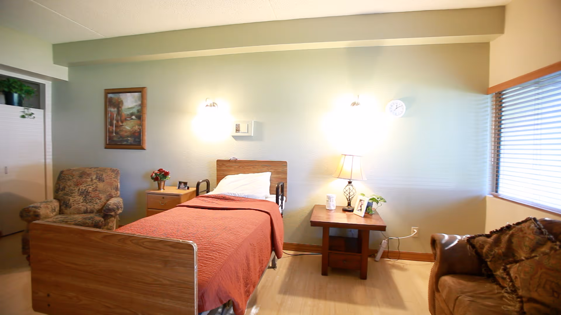 A cozy senior living bedroom with a single bed covered in a red blanket, a wooden headboard, and white pillow. There is a floral armchair to the left, a wooden nightstand with a vase of flowers and a picture frame, and a wooden side table with a lamp, a photo frame, and a small plant. The room has light-colored walls, a window with blinds on the right, and a framed painting on the wall above the armchair.