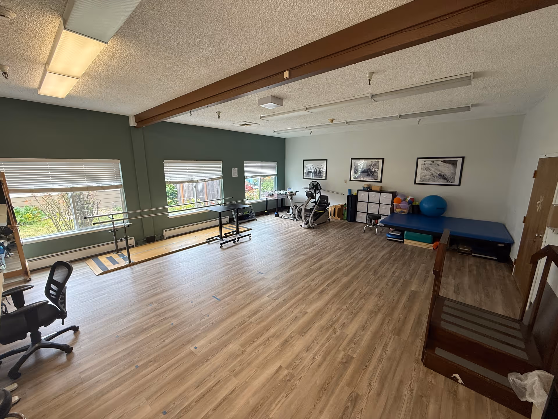 Large bright therapy/exercise room with parallel bars, exercise machines, a treatment table, and windows along one wall.