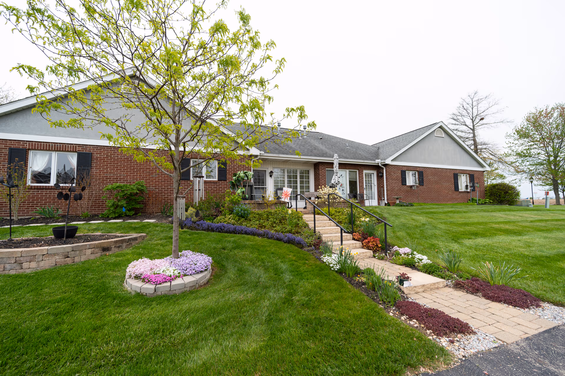 Single-story brick senior living building front with a paved walkway, steps with a handrail, and landscaped lawn and flower beds.