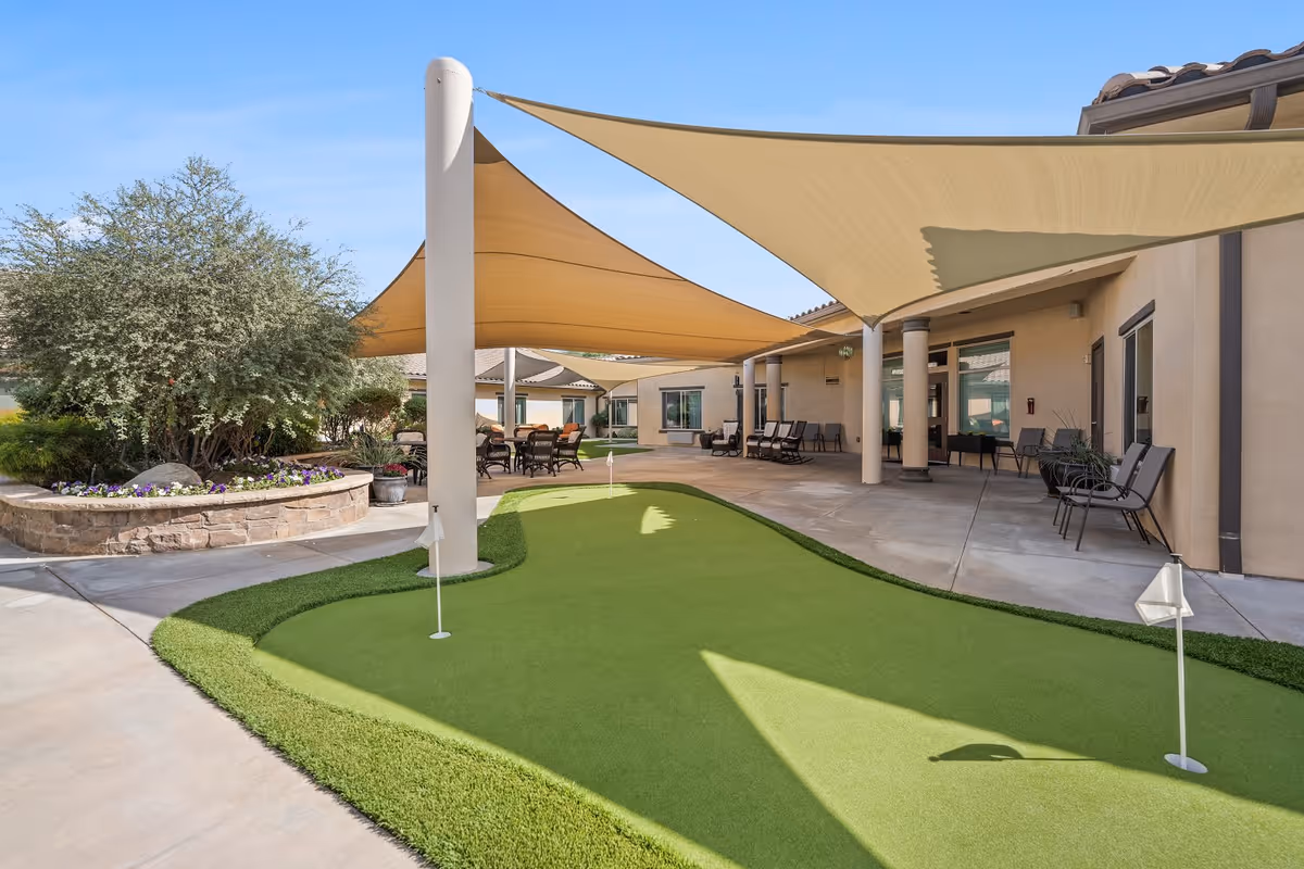 Outdoor courtyard area at Caleo Bay Assisted Living and Memory Care featuring a small putting green with flags, shaded seating areas with chairs and tables, and landscaping with bushes and flowers under a clear blue sky.