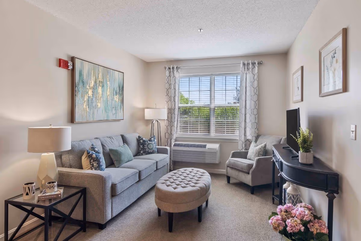 A cozy living room with a gray sofa adorned with decorative pillows, a matching armchair, and a tufted oval ottoman in the center. A large window with patterned curtains lets in natural light, and a black console table holds a flat-screen TV and a small plant. The walls are decorated with framed artwork, and a side table with a lamp and candles is placed next to the sofa.