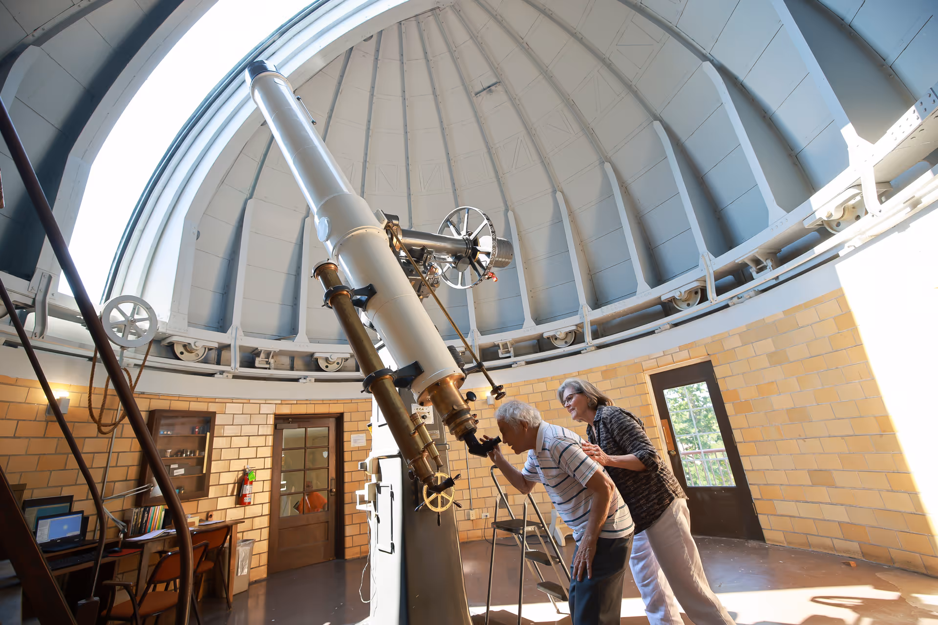 An elderly man looking through a large telescope inside an observatory dome, with a woman standing behind him offering support. The room has a high, curved ceiling with structural beams and yellow brick walls. There is a wooden door and a desk with a computer and books on the left side.