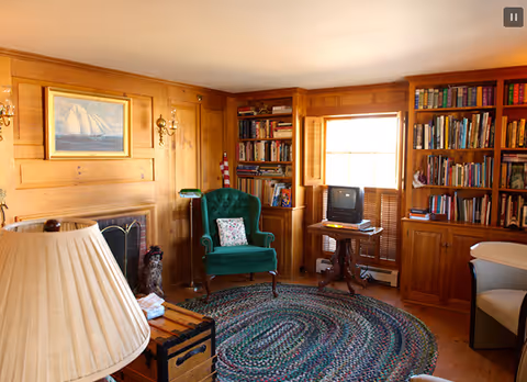 Cozy living room with wood-paneled walls, a green upholstered armchair, a round braided rug, built-in bookshelves filled with books, a small TV on a wooden table near a window with wooden shutters, and a fireplace with a painting of a sailboat above it.