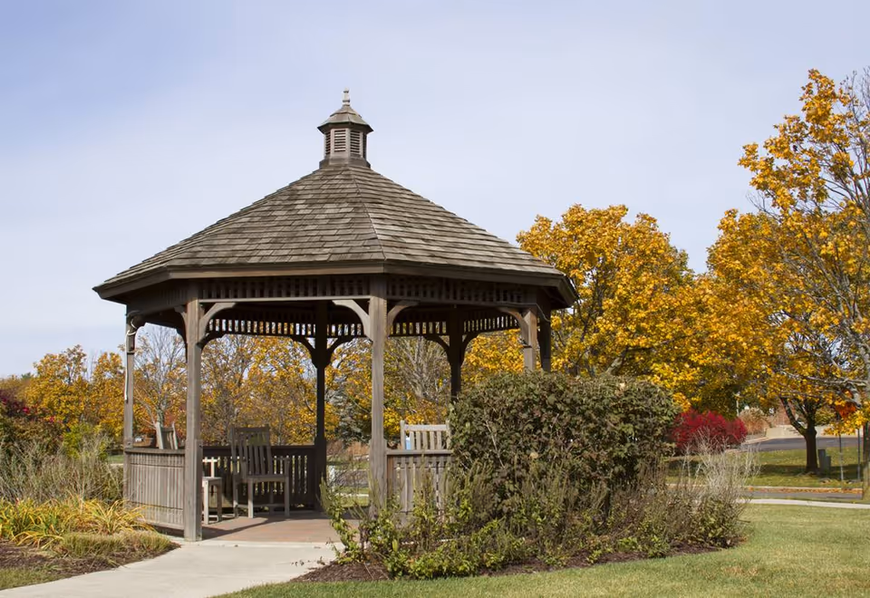 Wooden gazebo with benches set in a landscaped outdoor area with trees showing autumn foliage.