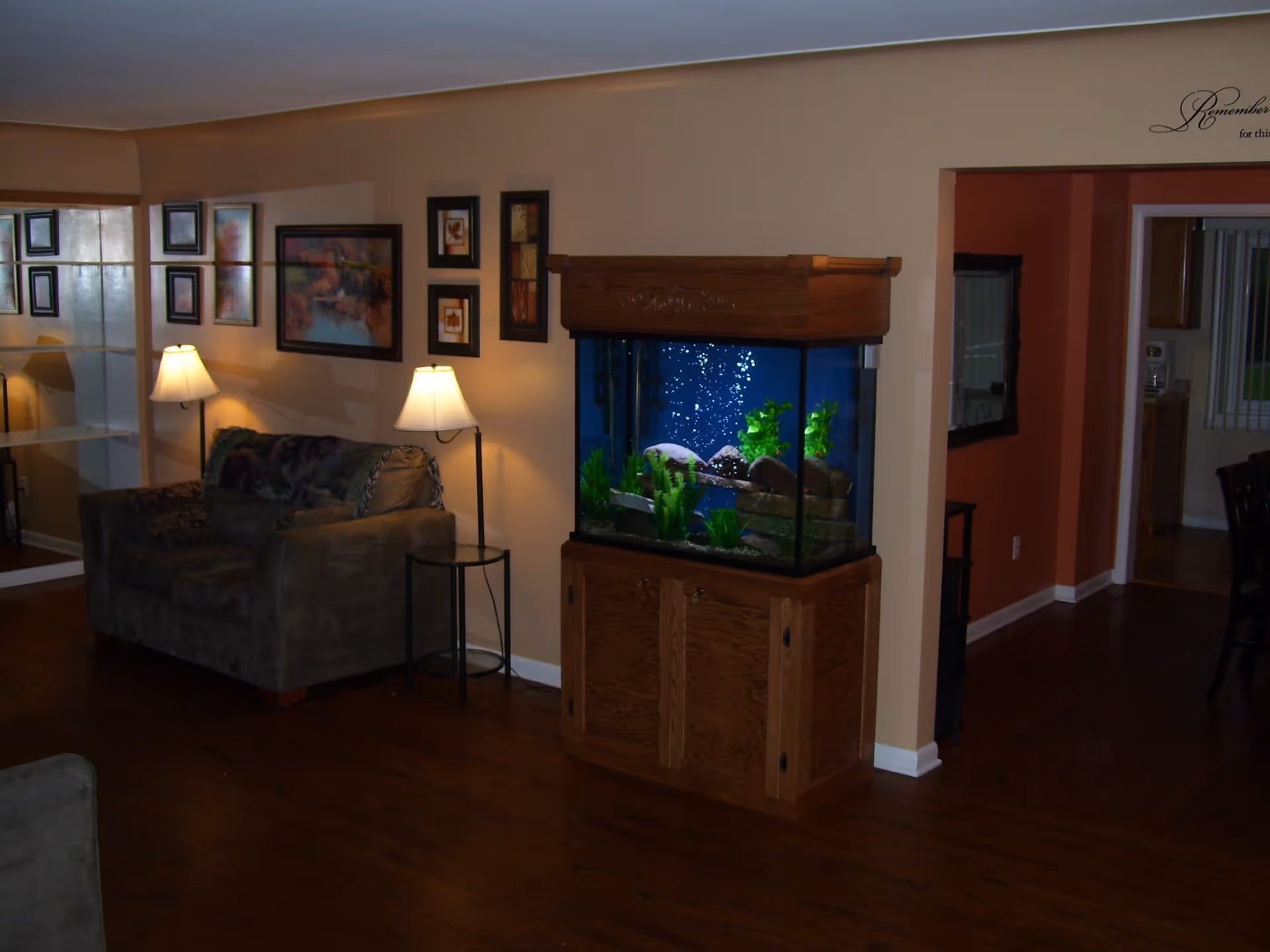 Interior of a living room with wooden flooring, a brown couch with a throw blanket, two floor lamps, several framed pictures on the wall, and a large fish tank with plants and rocks on a wooden cabinet. The room opens into another area with a red accent wall and a kitchen visible in the background.