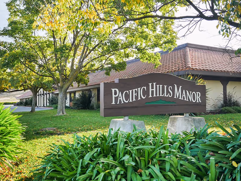 Outdoor view of Pacific Hills Manor facility sign surrounded by green plants and trees with a building featuring a tiled roof in the background.