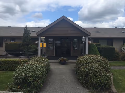 Front entrance of a single-story care facility with a peaked portico, glass doors, trimmed shrubs and a cloudy sky.