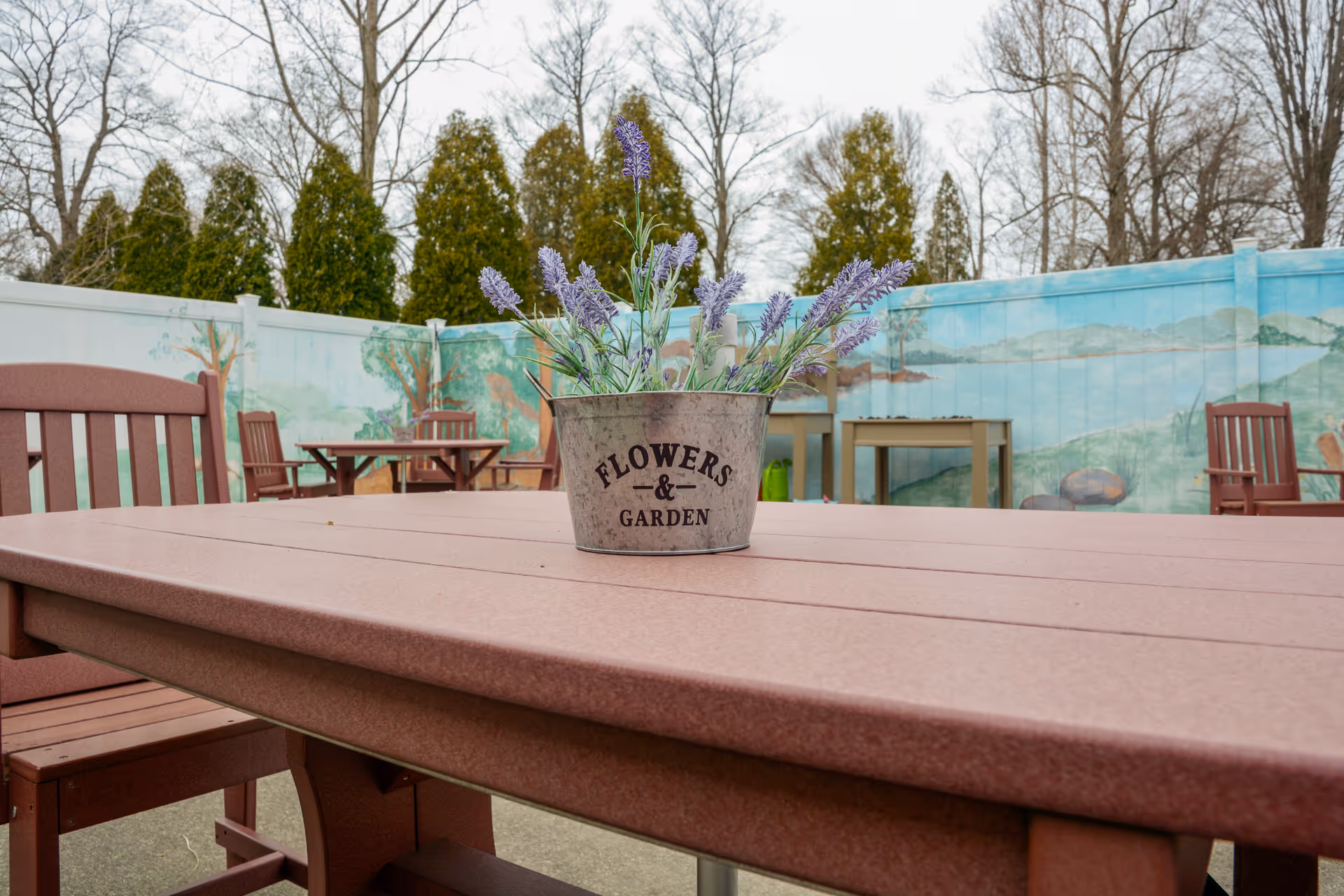 Outdoor patio area with brown wooden tables and chairs. A metal bucket labeled 'FLOWERS & GARDEN' containing purple flowers is placed on the table in the foreground. The background features a painted fence with nature scenes and tall evergreen trees behind it.