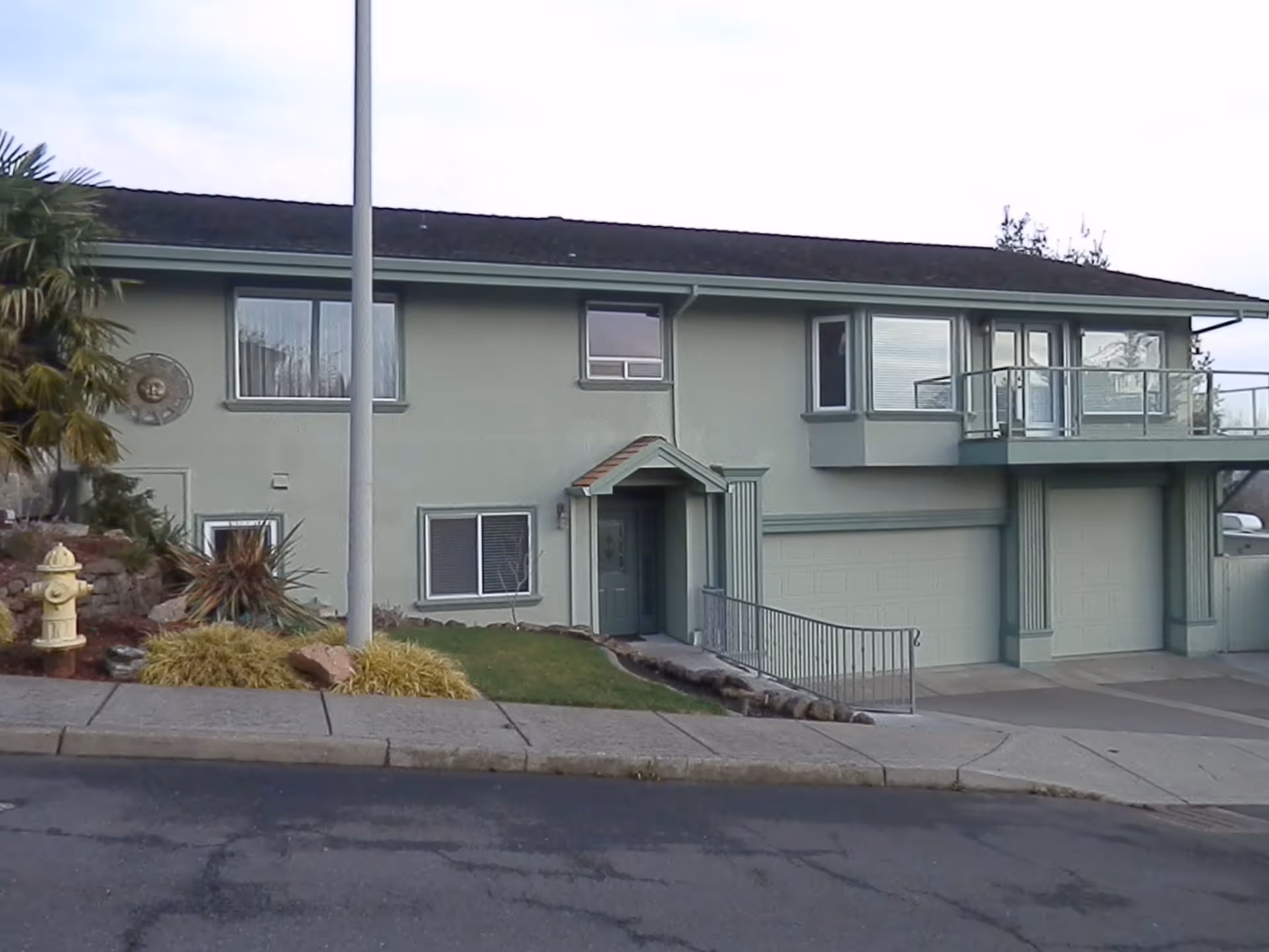 Exterior view of a two-story residential building with a light green facade, multiple windows, a garage with two doors, a small front porch with a slanted roof, a ramp with a railing, and landscaping including a palm tree and bushes. A yellow fire hydrant is visible on the left side near the sidewalk.