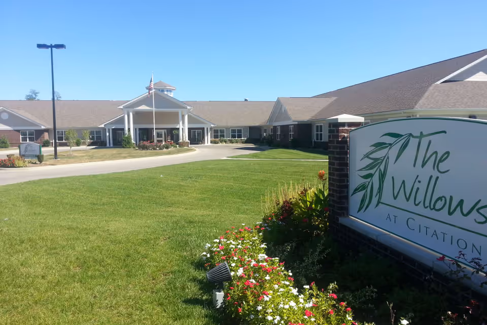 Front entrance of The Willows at Citation senior living facility with a circular drive, manicured lawn, flower beds, and the facility sign in the foreground.