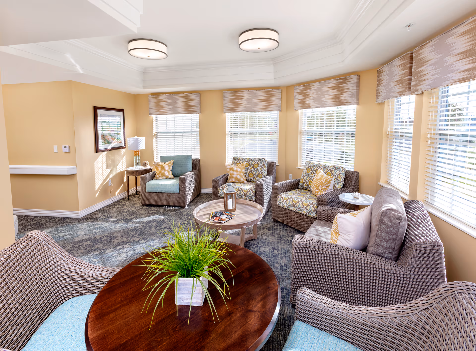Sunlit common sitting room with wicker armchairs, a round wooden table topped by a potted plant, and multiple windows with blinds.