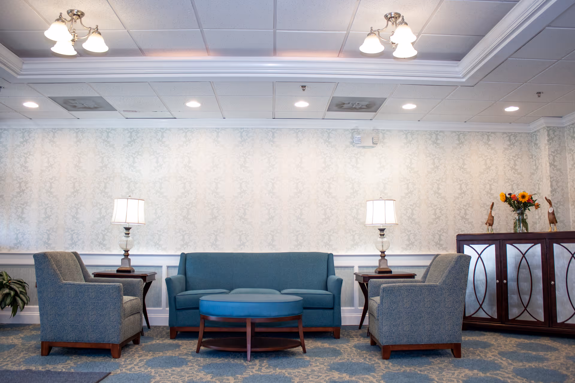 A well-lit sitting area with a blue upholstered sofa and two matching armchairs arranged around a round blue ottoman. There are two side tables with lamps on either side of the sofa. A wooden cabinet with decorative items and a vase of flowers is positioned against the wall. The room has patterned wallpaper, a carpeted floor, and ceiling lights.