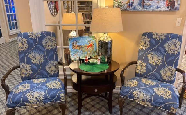 Two blue patterned armchairs flank a round wooden side table topped with a lamp and holiday decorations in a carpeted seating area.