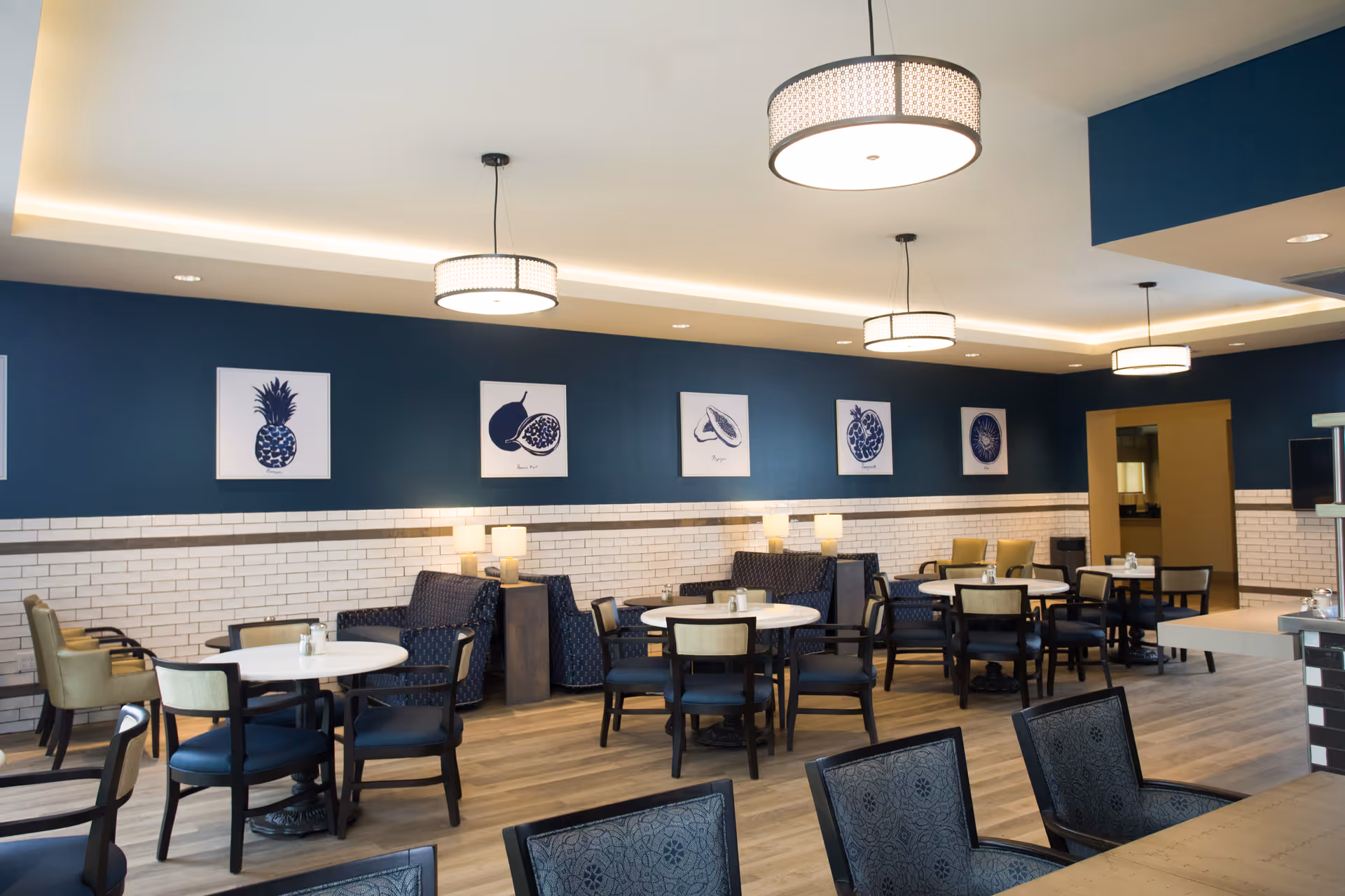 A dining room with multiple round tables and chairs arranged neatly. The walls are painted dark blue with white subway tile halfway up. There are framed fruit illustrations hanging on the wall. Modern pendant lights hang from the ceiling, and the floor is wood. The room has a clean, contemporary look.