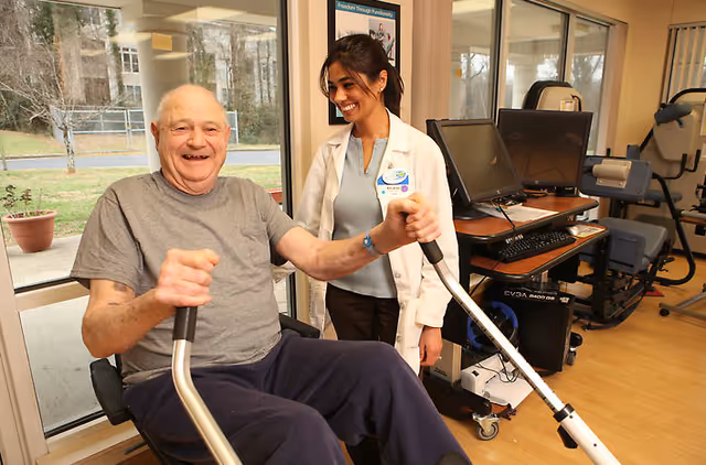 An elderly man is exercising on a seated rowing machine in a fitness or rehabilitation room. A female healthcare professional in a white coat stands beside him, smiling and offering support. The room has large windows showing an outdoor view, exercise equipment, and computer monitors in the background.
