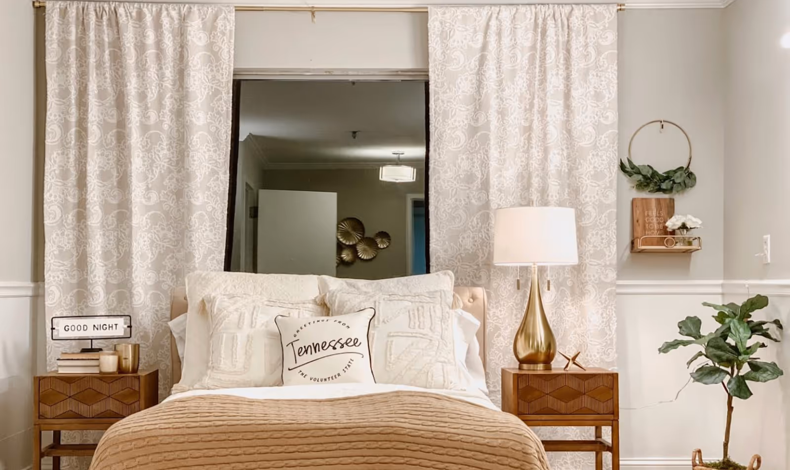 A cozy bedroom with a neatly made bed featuring cream and beige bedding. The bed has decorative pillows, including one with the text 'Greetings from Tennessee The Volunteer State.' On either side of the bed are wooden nightstands; the left one has a small sign that says 'GOOD NIGHT' along with books and candles, and the right one has a gold lamp and a small decorative item. Behind the bed are patterned curtains, and to the right on the wall is a small shelf with a plant and a sign that reads 'FEELS GOOD TO BE HOME.' A potted plant is placed on the floor next to the nightstand.