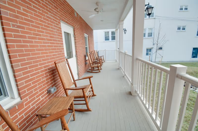 A covered outdoor porch area with several wooden rocking chairs lined up along a red brick wall. The porch has white railings and a ceiling fan, overlooking a grassy area and another building in the background.