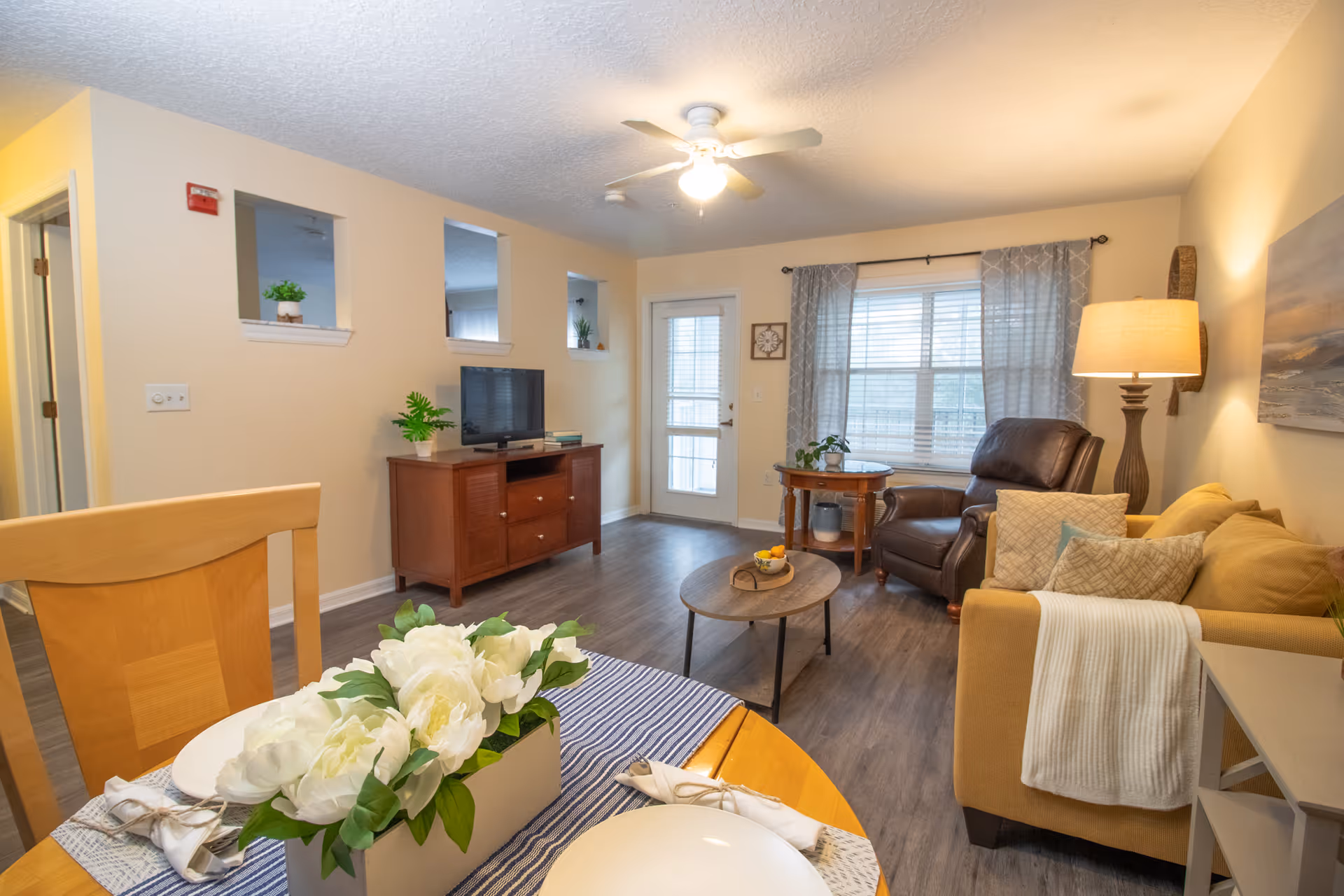 Sunny furnished living room with a sofa and armchair, coffee table, TV stand, dining table with flowers, and a door leading outside.