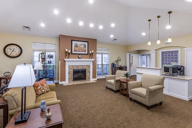 A spacious living room area in a senior living facility with beige walls and carpet. The room features a fireplace with a framed picture above it, two windows on either side, a clock on the wall, and several pieces of furniture including a sofa, two armchairs, a side table with a lamp, and a reception desk with pendant lights hanging above.
