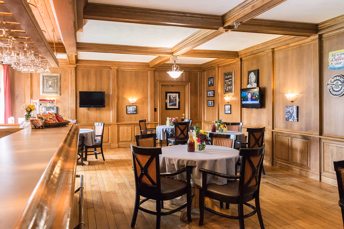Wood-paneled dining room with round tables covered in white tablecloths, chairs, floral centerpieces, and a small bar area.