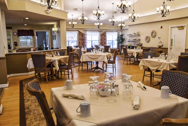 Dining room with multiple set tables and chairs, chandeliers, and large windows in an assisted living facility.