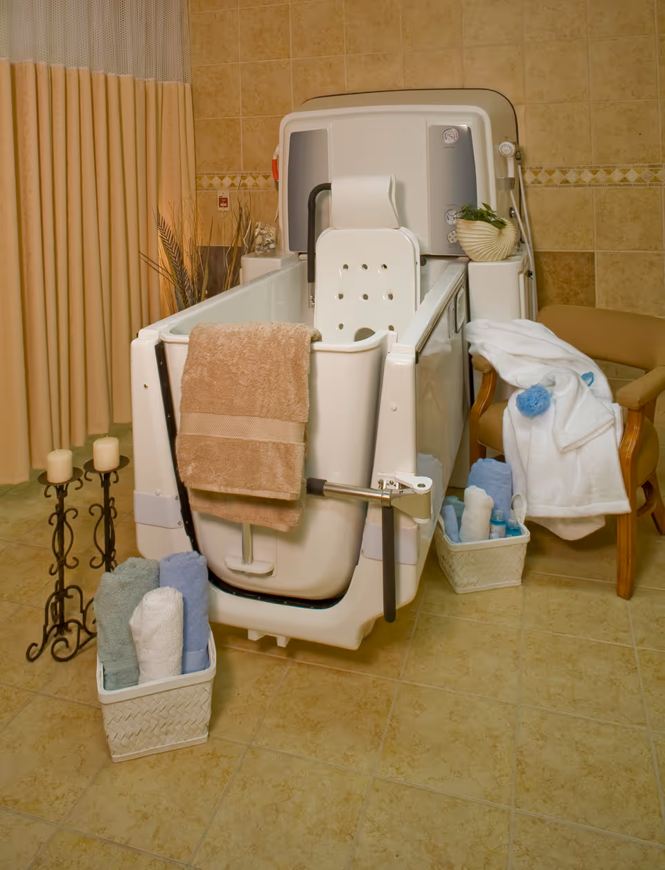 A walk-in bathtub with a white seat and safety handles in a tiled bathroom. A beige towel hangs over the tub door. Nearby are baskets with rolled towels and toiletries, a beige armchair with a white robe draped over it, and a decorative candle holder with three candles. The room has beige tiled walls and floor with a curtain on one side.