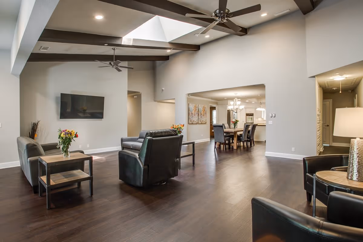 Spacious living room area with dark wood flooring, leather armchairs and sofas, a wall-mounted flat screen TV, ceiling fans, and a dining area with a wooden table and chairs in the background. The room has high ceilings with exposed beams and a skylight.