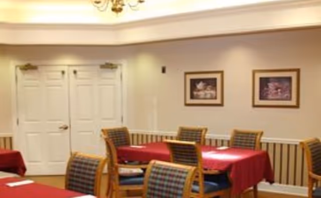 Interior view of a dining room with tables covered in red tablecloths and wooden chairs with patterned cushions. Two framed pictures hang on the beige wall, and there are double white doors in the background.