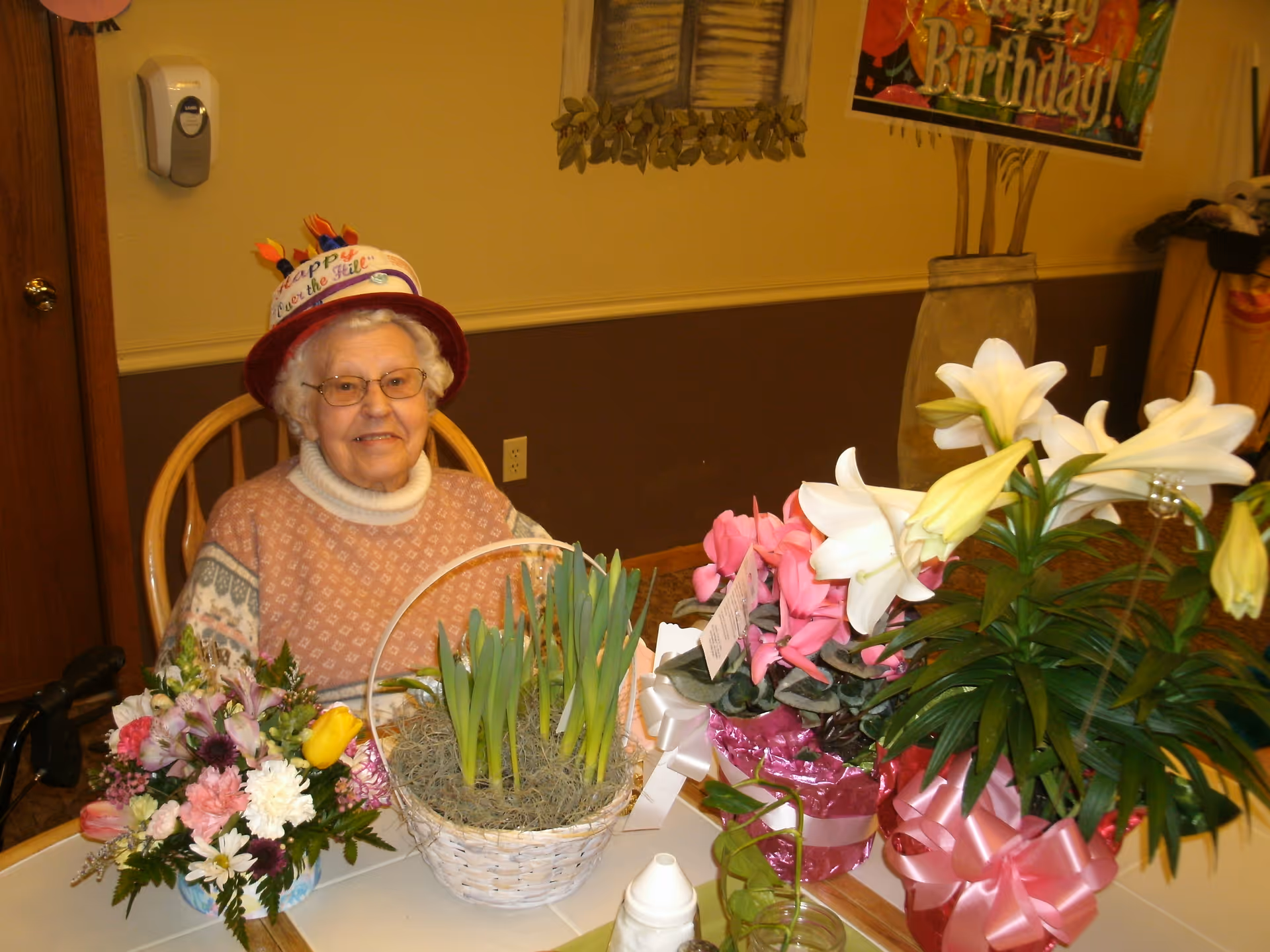 An elderly woman wearing a birthday hat sits at a table surrounded by potted flowers and bouquets.