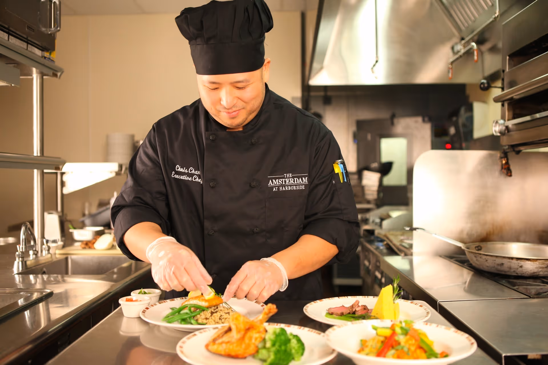 A chef wearing a black uniform and hat is preparing plated meals in a commercial kitchen. The chef is arranging food on a plate with green beans and rice, with other plates of food including chicken, broccoli, and a dish with vegetables visible on the counter.