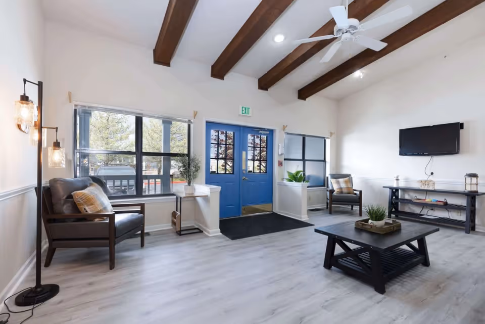Bright lobby/common area with blue double doors, seating, a coffee table, wall-mounted TV and exposed ceiling beams.