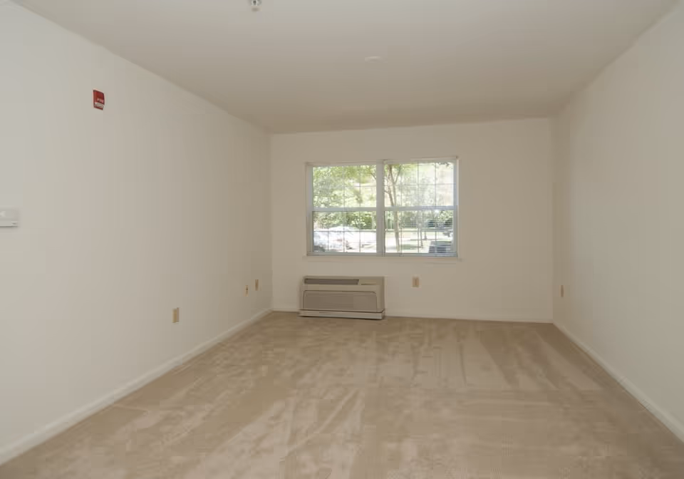 Empty carpeted room with white walls, a window showing trees outside, and an HVAC unit beneath the window.