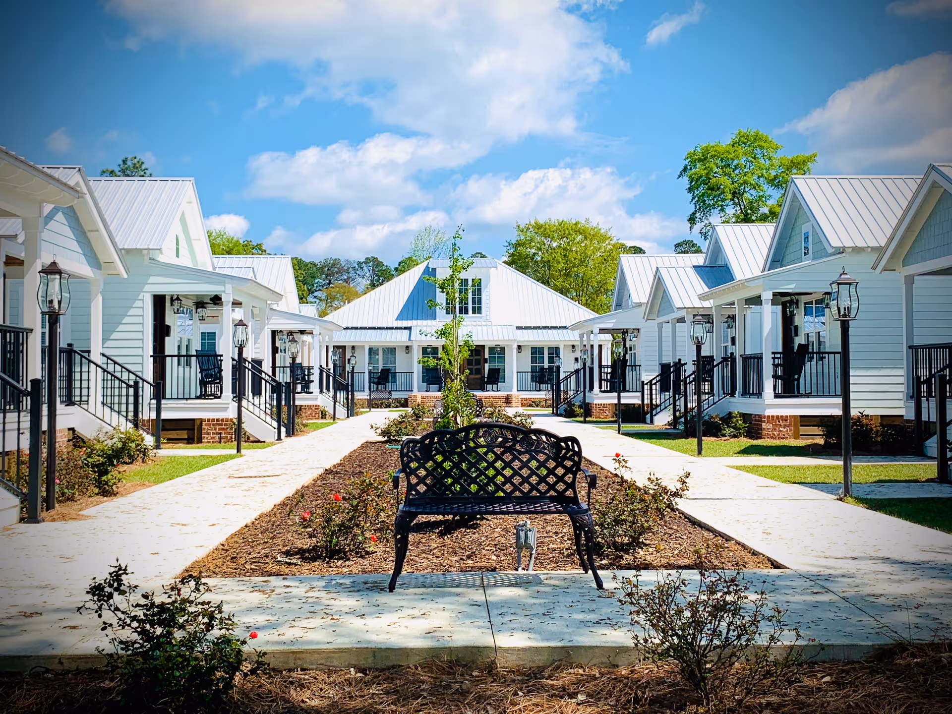 A peaceful outdoor courtyard area at James Place Senior Living Residents featuring a black metal bench in the center, surrounded by landscaped flower beds and small bushes. The courtyard is flanked by white cottages with metal roofs, each having a porch with black railings and outdoor lamps. The sky is bright blue with scattered clouds.