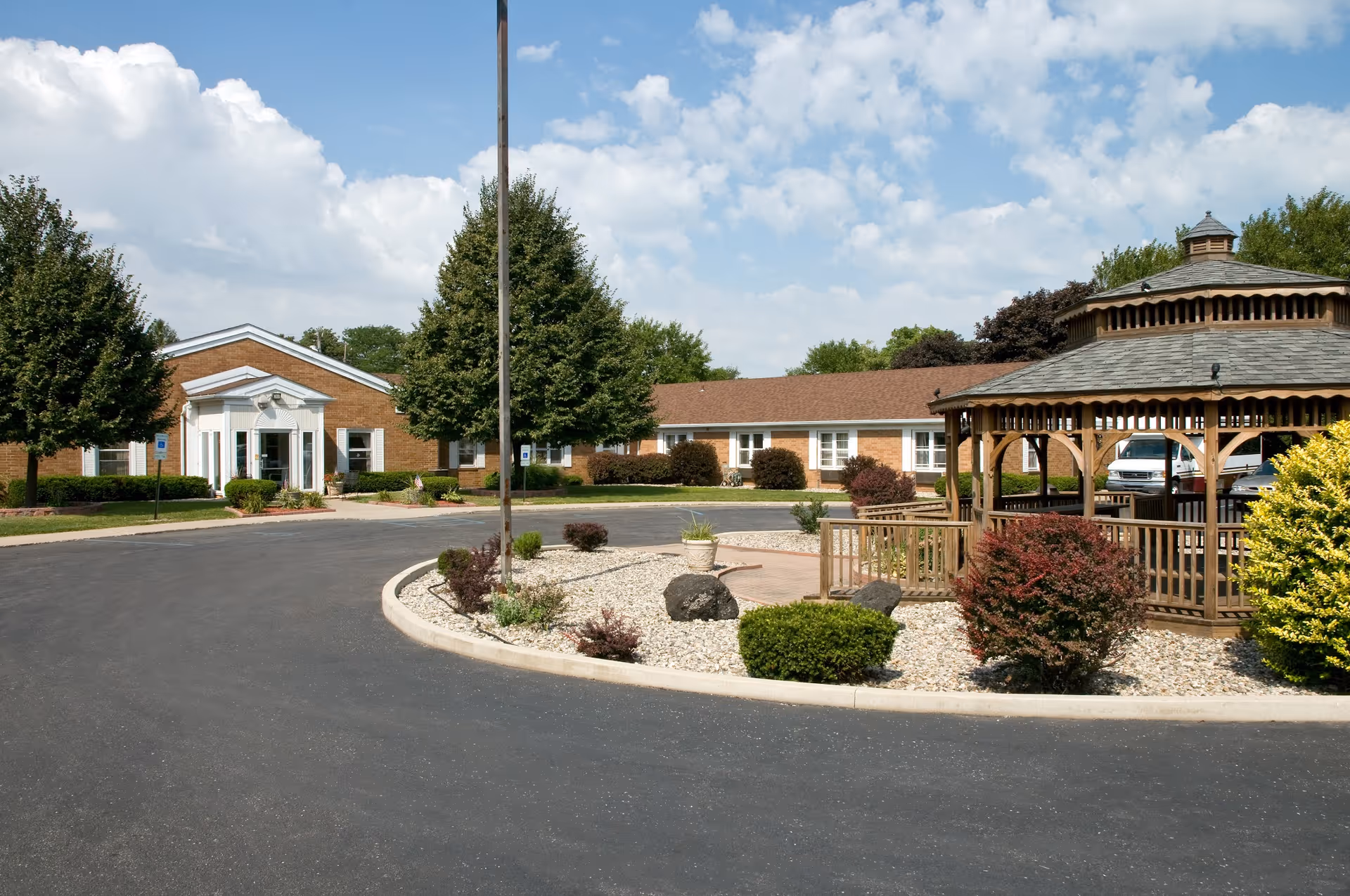 Front entrance of a single-story brick care center with a circular driveway, landscaped rock island, and a wooden gazebo.