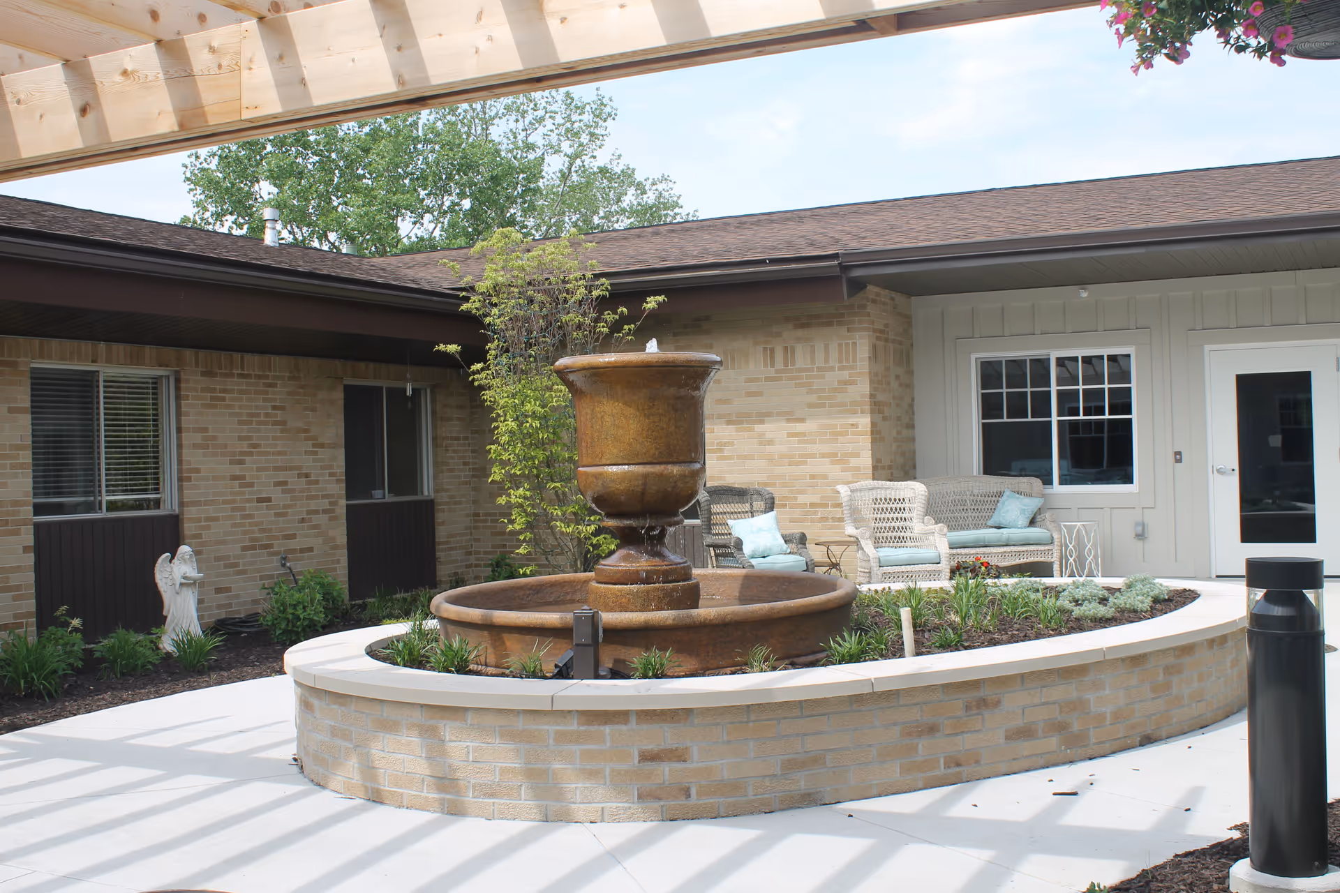 Outdoor courtyard area with a large circular brick planter featuring a central water fountain. Surrounding the planter are some green plants and shrubs. There are wicker chairs with cushions and a small table near the building wall. The building has brick and light-colored siding with windows and a door. A wooden pergola casts shadows on the ground.