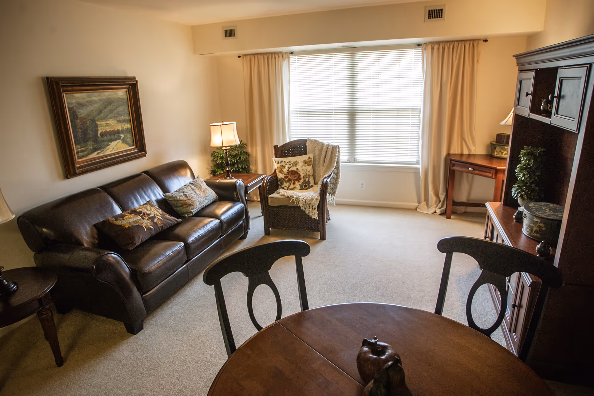 A cozy living room with a dark brown leather sofa adorned with decorative pillows, a wicker armchair with a floral cushion and throw blanket, a wooden round dining table with chairs, a wooden cabinet with plants and decorative items, a side table with a lamp, and a large window with beige curtains letting in natural light.