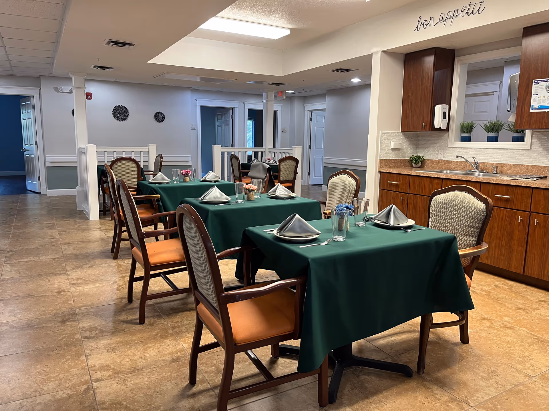 Dining area with tables covered in green tablecloths set with napkins and glassware, and a kitchenette in the background under a "bon appetit" sign.