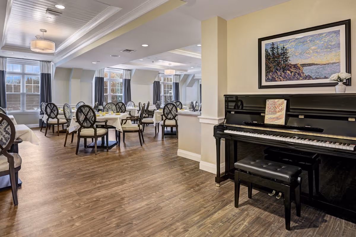 Bright communal dining room with multiple round tables and chairs and an upright piano against a wall.