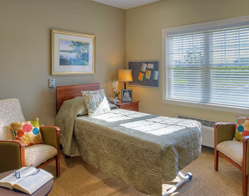 Sunlit private bedroom with a single bed, two armchairs, a nightstand with a lamp, a window with blinds, and framed wall art.
