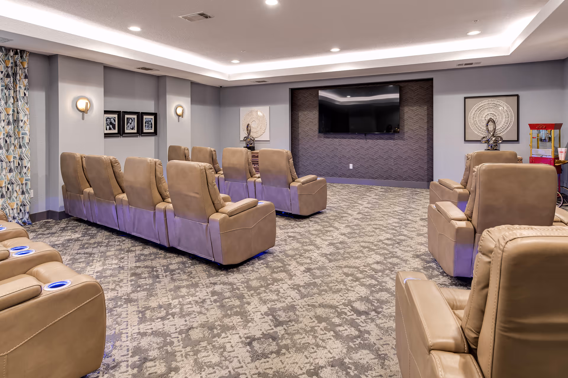 A cozy media room with multiple rows of beige leather recliner chairs facing a large flat-screen TV mounted on a textured dark accent wall. The room has patterned carpet flooring, soft wall lighting, framed artwork, and a popcorn machine in the corner.