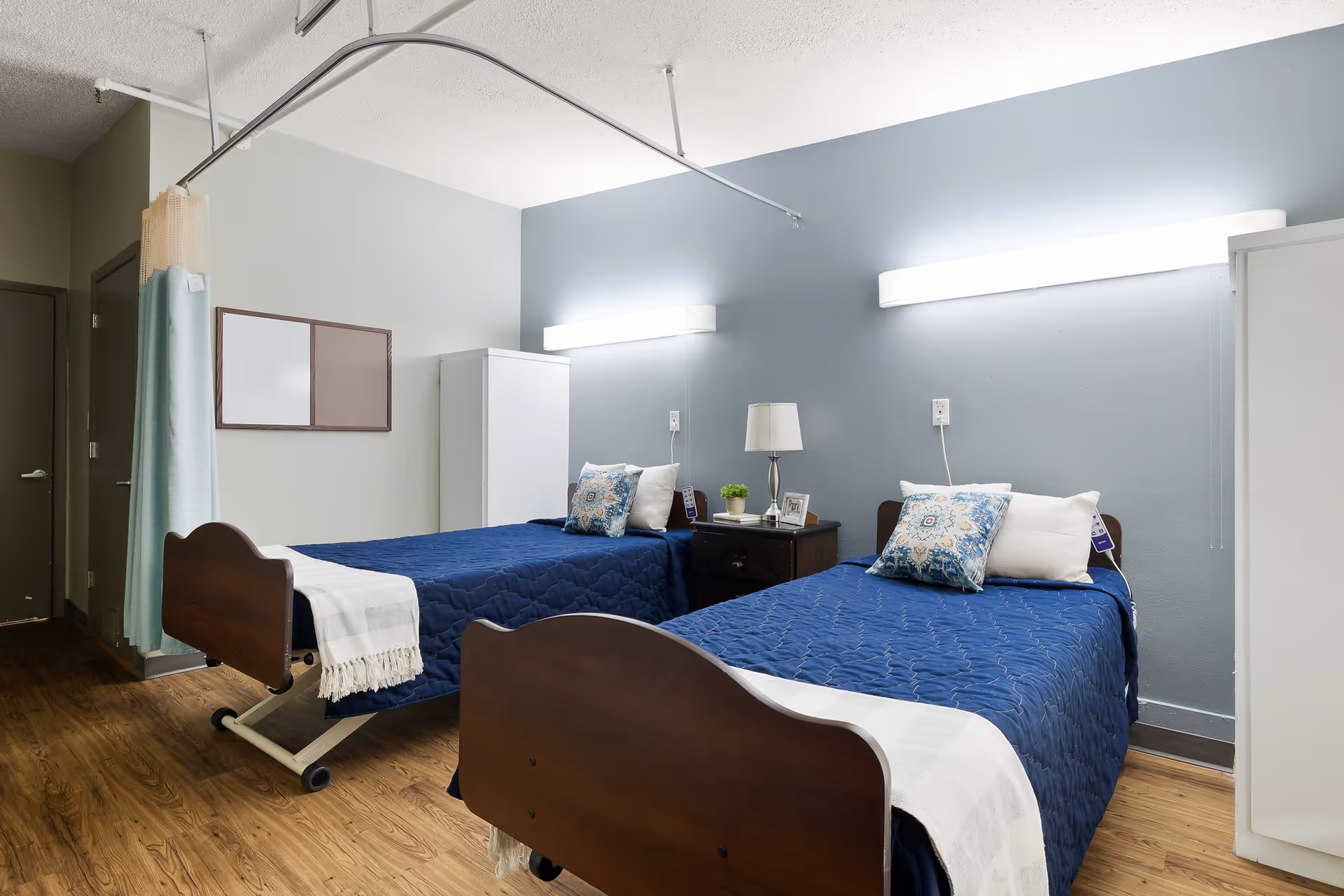 A nursing facility bedroom with two single beds covered in blue quilts and white pillows. Between the beds is a dark wooden nightstand with a lamp, a small plant, and picture frames. The room has light blue and gray walls, wooden flooring, a white wardrobe, and a privacy curtain on a ceiling track.