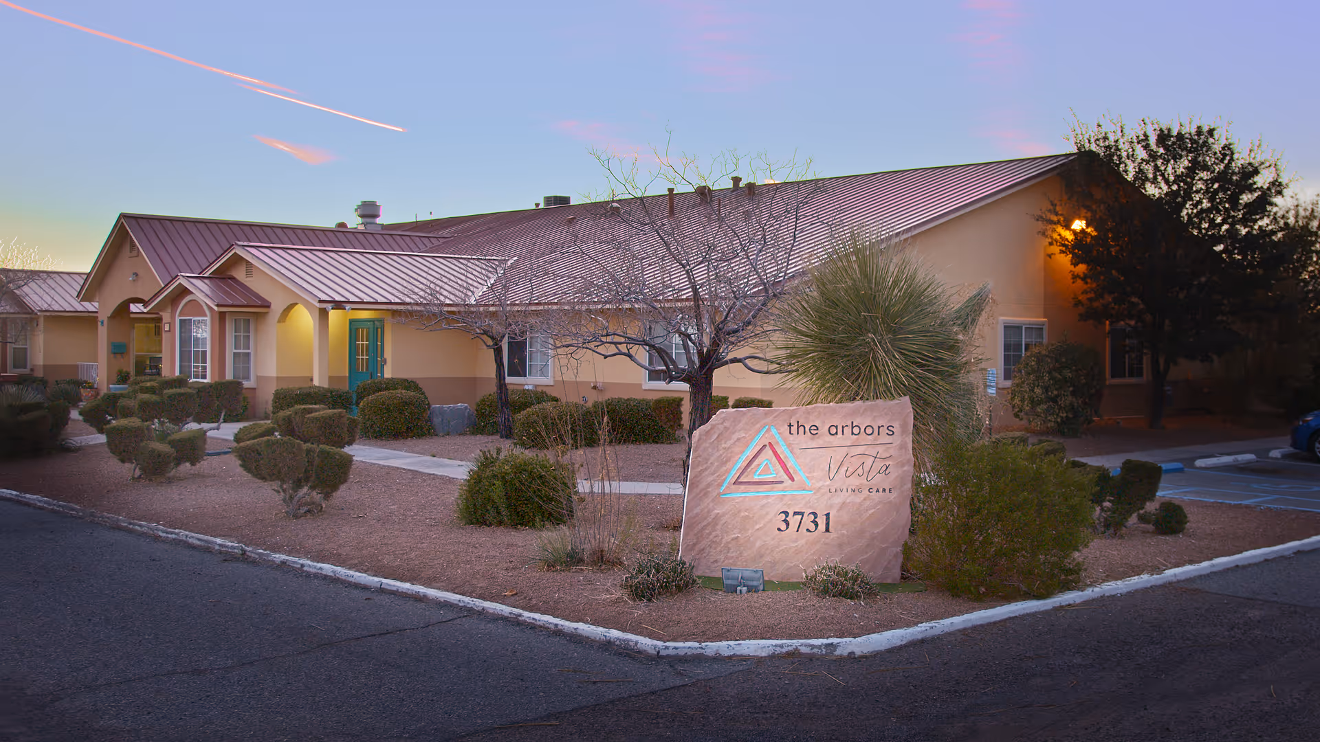 Exterior view of The Arbors at Vista Living Care facility at dusk, showing a single-story building with a reddish-brown roof, beige walls, and a landscaped front yard with trimmed bushes and trees. A large stone sign with the facility's name and address (3731) is prominently displayed in the foreground.