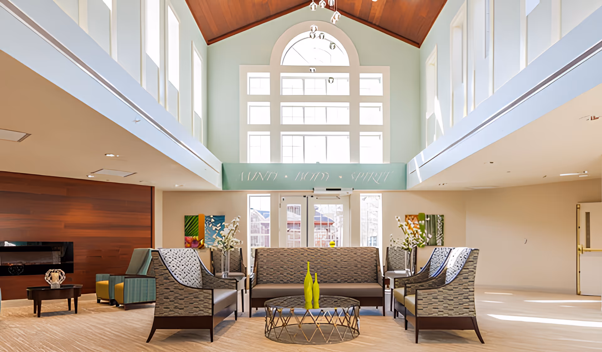 A spacious and well-lit living room area with high ceilings and large windows allowing natural light to fill the space. The room features a set of patterned armchairs and sofas arranged around a circular glass coffee table with decorative green vases. The walls are light-colored with some colorful artwork and floral arrangements on side tables. There is a wooden accent wall with a fireplace on the left side.