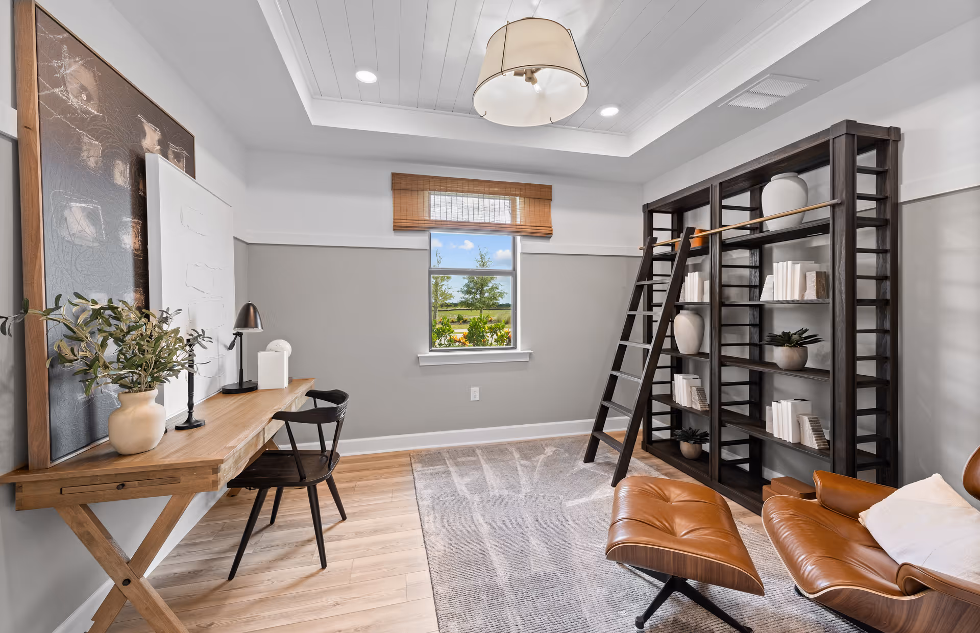 A cozy interior room featuring a wooden desk with a chair, a table lamp, and decorative items including a plant and artwork. There is a large dark wooden bookshelf with a ladder, filled with books and vases. A comfortable brown leather lounge chair with an ottoman is positioned on a gray patterned rug. A window with a bamboo shade lets in natural light, and a ceiling light fixture hangs above.
