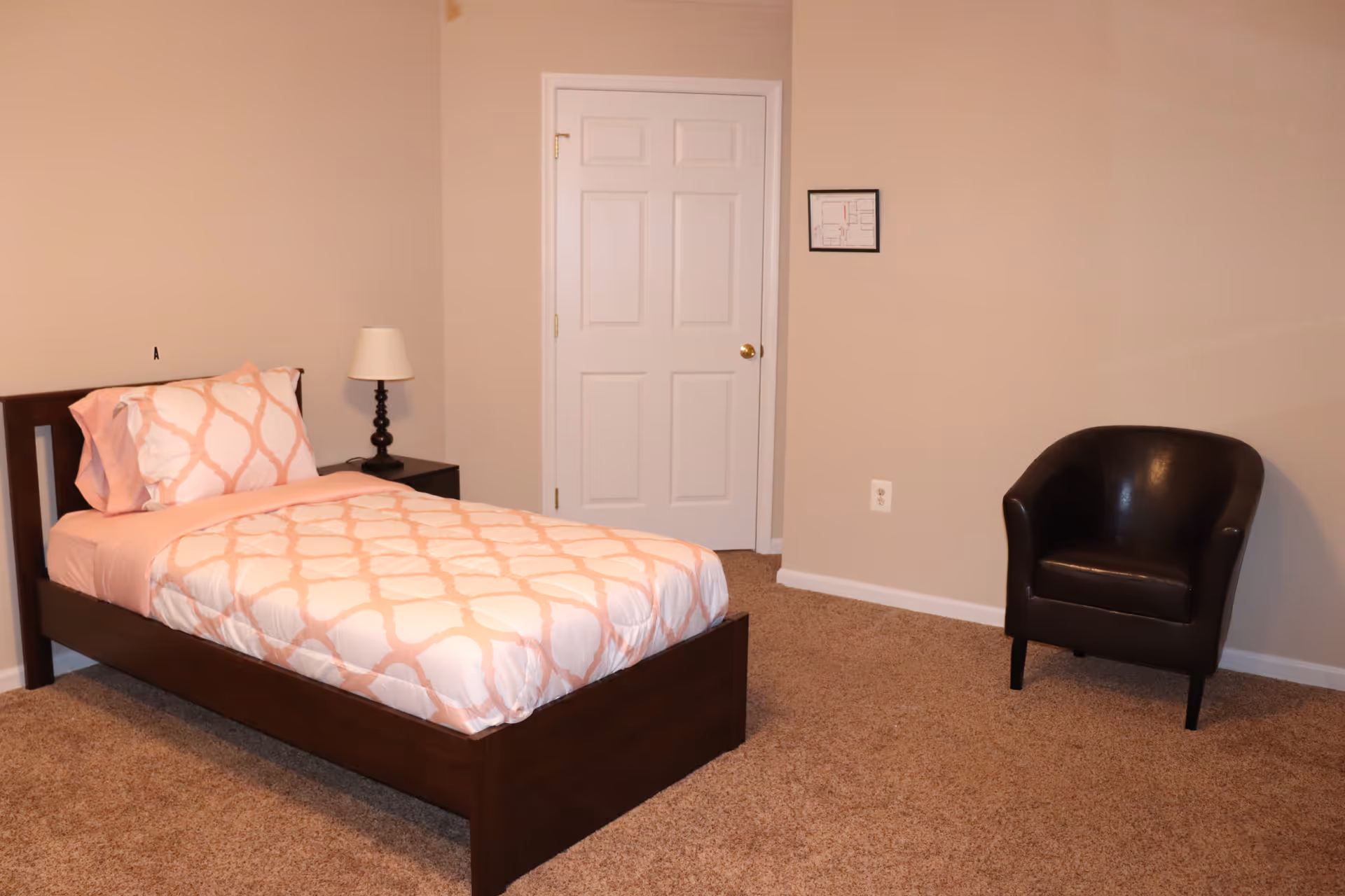 A simple bedroom with a single bed featuring a pink and white patterned comforter and pillow, a dark wooden nightstand with a lamp, a closed white door, a small framed floor plan on the beige wall, and a dark brown armchair on a carpeted floor.