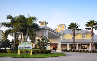 Exterior view of The Brennity at Tradition Senior Living facility showing a large building with a covered entrance, palm trees, and a clear blue sky.