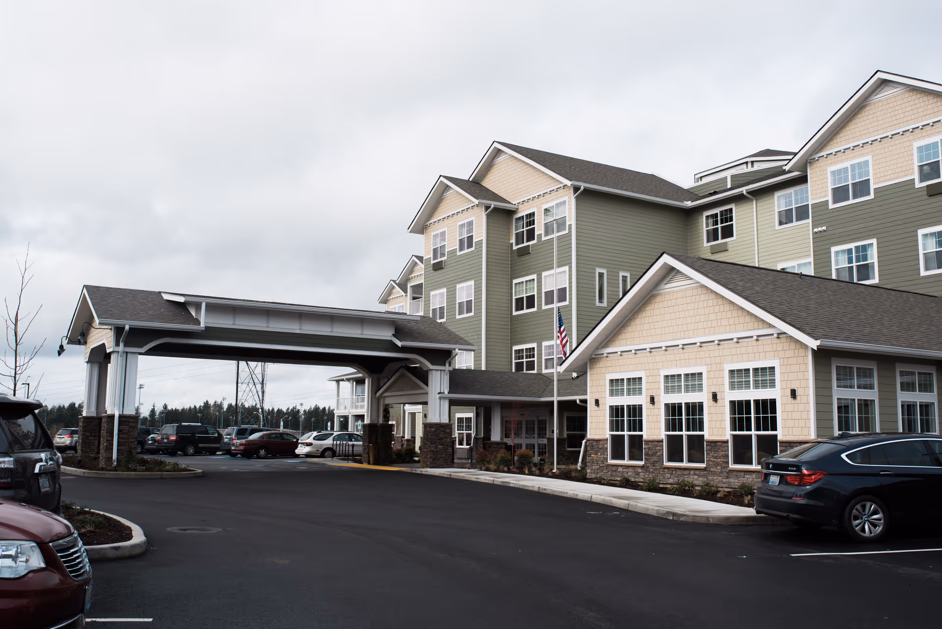 Exterior view of a multi-story senior living facility building with green and beige siding, multiple windows, and a covered entrance driveway. Several cars are parked in the parking lot under a cloudy sky.