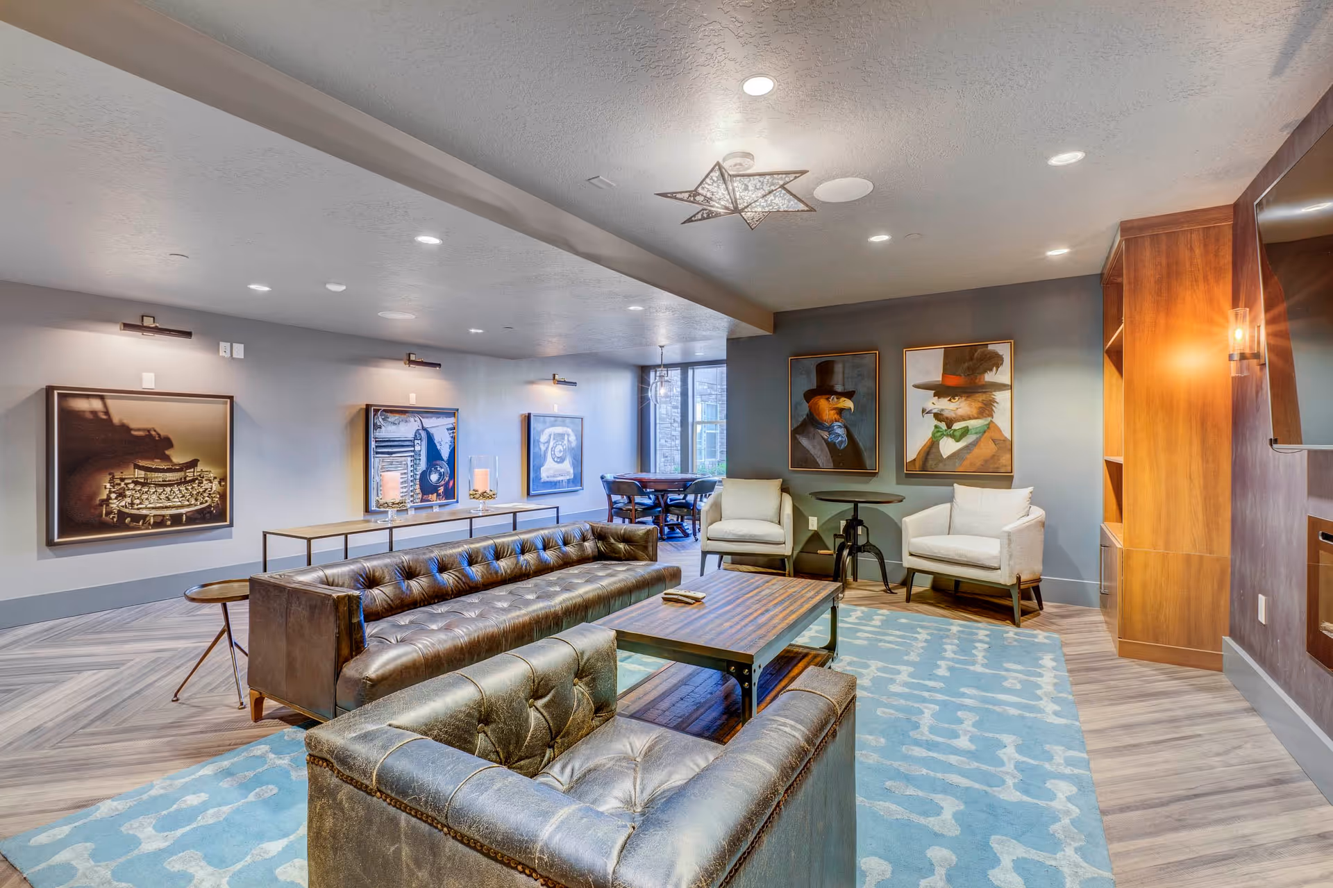 A modern living room area with two dark leather tufted sofas facing a wooden coffee table on a blue patterned rug. The walls are decorated with framed artwork, including two paintings of animals dressed in formal attire. There are two white armchairs with a small round table between them, and a large flat-screen TV mounted on a wooden panel on the right wall. The room has recessed lighting and a star-shaped ceiling light fixture.
